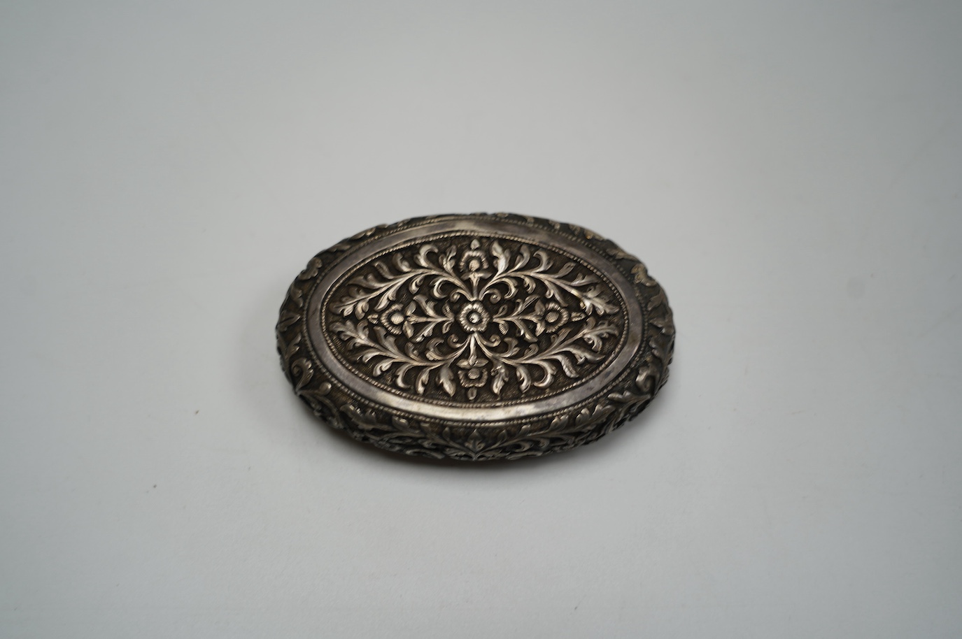 An Indian embossed white metal oval tobacco box, 97mm. Condition - poor to fair